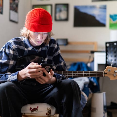 Junge mit rotem Beany hält eine Gitarre und das Smartphone in der Hand