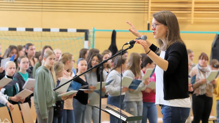 Chorleiterin Susanne Blumenthal vor einem Mikrofon. Schülerinnen und Schüler unscharf im Hintergrund sichtbar (Foto: SWR, Johannes Riedel)