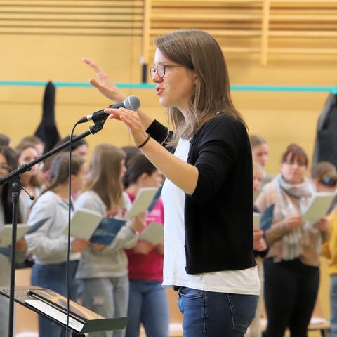 Chorleiterin Susanne Blumenthal vor einem Mikrofon. Schülerinnen und Schüler unscharf im Hintergrund sichtbar (Foto: SWR, Johannes Riedel)