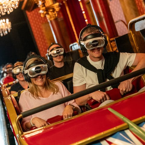 Vollbesetzer Achterbahn-Waggon; die Menschen haben Kopfhörer und Virtual-Reality-Brillen auf, im Hintergrund sieht man Kronleuchter und rot gemusterte Tapeten.