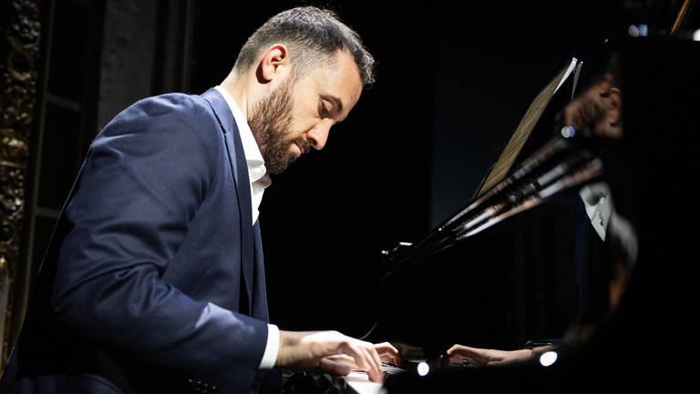 Igor Levit spielt im dunkelblauem Anzug vor schwarzem Hintergrund am Klavierflügel