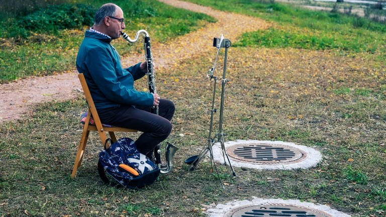 Mann auf Klappstuhl spielt Bassklarinette im Freien vor einem Gully (Foto: SWR, Ralf Brunner)