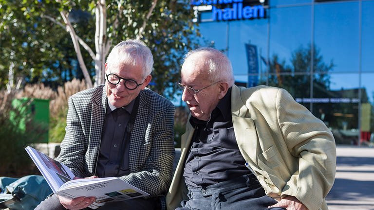 Zwei Männder mit Brillen sitzen auf einer Bank und schauen in ein Buch