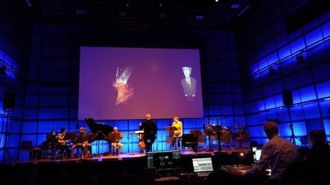 Ensemble mit Dirigent und Sängerin auf Bühne mit Leinwand im Hintergrund (Foto: SWR)