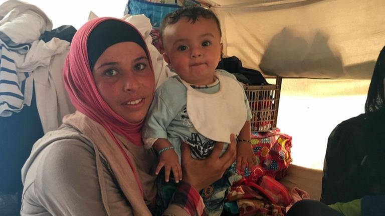 Eine junge Frau mit einem Kleinkind auf dem Arm sitzt in einem Zelt in einem Flüchtlingslager. Beide schauen in die Kamera. (Foto: SWR, SWR - Anna Osius)
