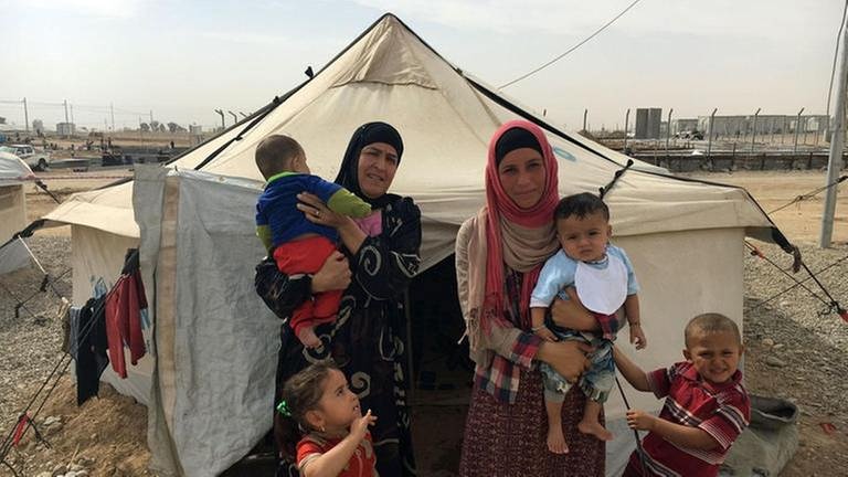 Zwei Frauen mit Kopftuch stehen vor einem Zelt in einem Flüchtlingslager. Sie halten zwei Kleinkinder im Arm. Zwei etwas größere Kinder stehen neben ihnen. (Foto: SWR, SWR - Anna Osius)