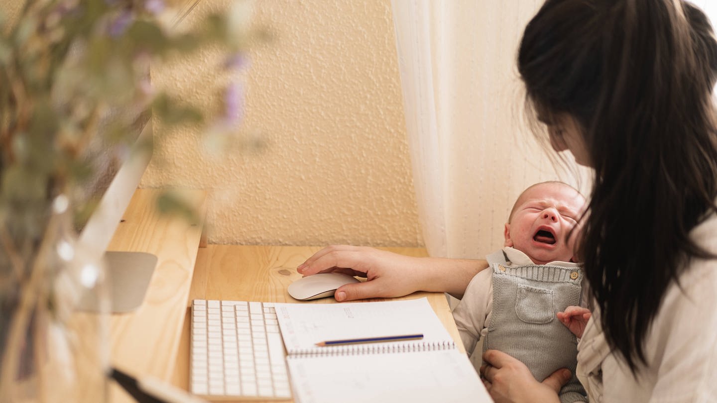 Eine Frau arbeitet am Computer und hat dabei ihr Baby auf dem Arm (Foto: IMAGO, IMAGO / Addictive Stock)