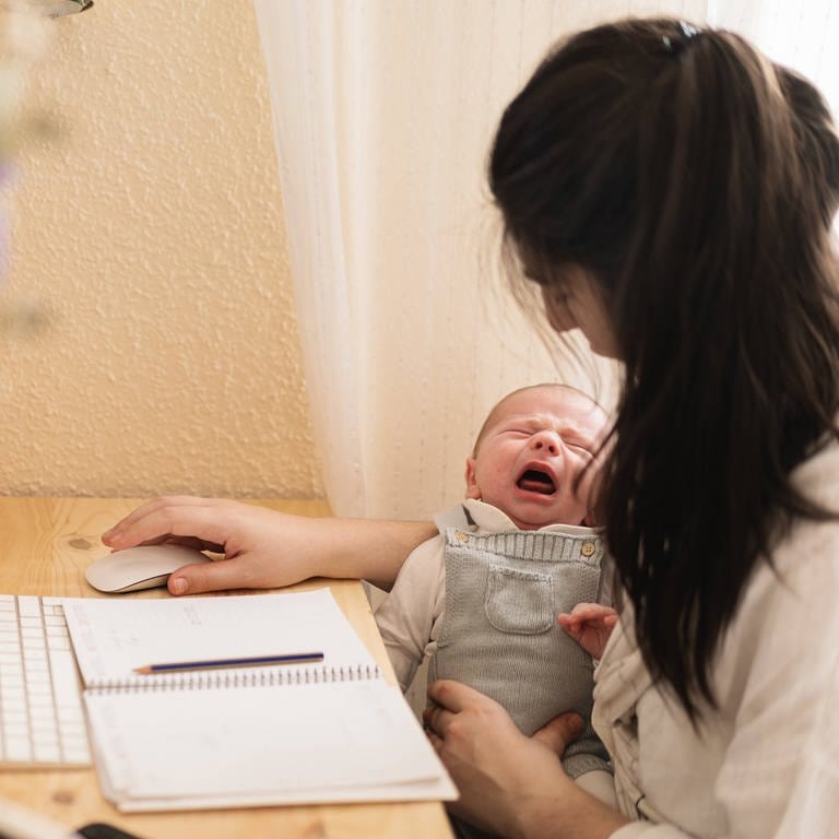 Eine Frau arbeitet am Computer und hat dabei ihr Baby auf dem Arm (Foto: IMAGO, IMAGO / Addictive Stock)