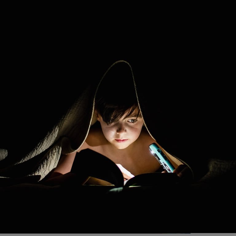 Ein Kind liegt im Bett und liest unter der Decke ein Buch bei Nacht (Foto: IMAGO, IMAGO / Cavan Images)