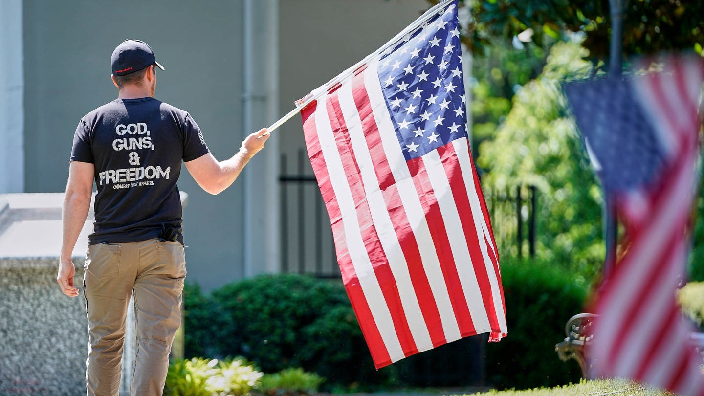God, Guns & Freedom: Ein Mann mit der US-Flagge bei Protesten am 2. Mai 2020 in Morganton / North Caroline. Gut 50 Menschen forderten den Gouverneur von North Carolina, Roy Cooper, auf, die Beschränkungen, die wegen der COVID-19-Pandemie verhängt worden waren, zu lockern. (Foto: IMAGO, imago images / ZUMA Wire)
