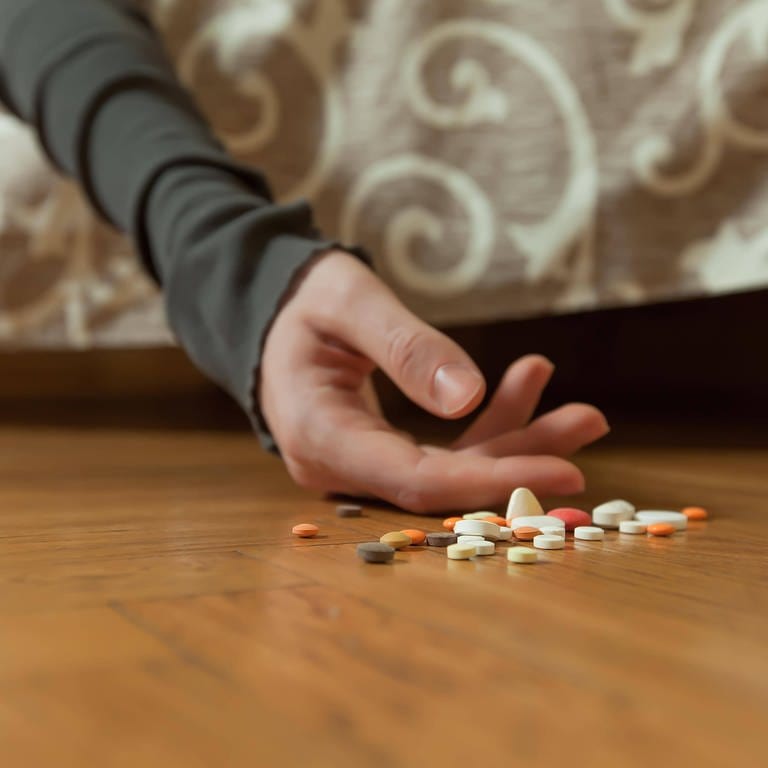 Eine Hand hängt von einem Bett, daneben Tabletten: 10.000 Selbtmorde pro Jahr in Deutschland: Durch gezielte Schulung von Ärzten, aber auch Aufklärung der Gesamtbevölkerung wollen Forscher suizidgefährdete Menschen besser unterstützen. 