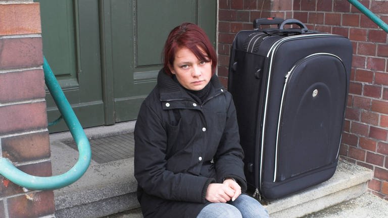 Junge Frau sitzt allein mit Koffer auf einer Treppe: Mit 18 Jahren erwachsen – für Pflege- und Heimkinder endet dann in der Regel die Jugendhilfe. Die jungen Menschen stehen plötzlich alleine da und werden zu Niemandskindern: Niemand fühlt sich für sie verantwortlich – auch die Behörden nicht.