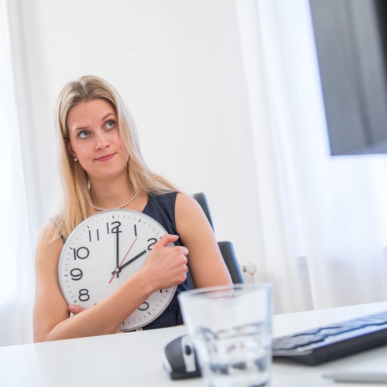 Frau hält am Schreibtisch große Uhr: Mütter und Väter haben ein Recht, nach der Elternzeit tageweise oder halbtags zu arbeiten – so manches Mal landen sie in Teilzeit aber nur auf unattraktiven Positionen.