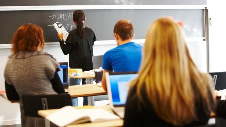 Jugendliche sitzen in einem Unterrichtsraum. Eine Lehrkraft schreibt mit Kreide an eine Tafel. (Foto: Colourbox, Model Foto: Colourbox.de -)
