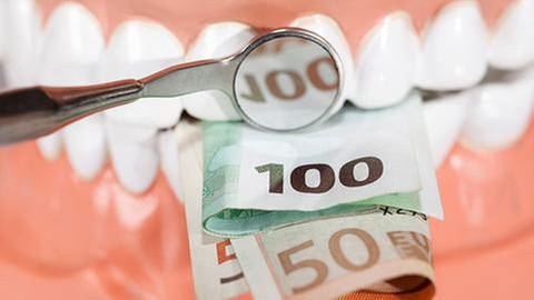 Ein Zahnarztspiegel beleuchtet Geldscheine, die in einem Gebiss stecken. (Foto: Getty Images, Thinkstock -)