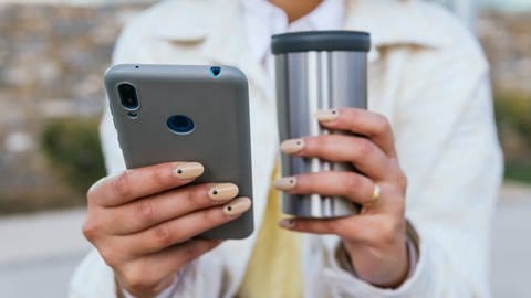 Frau mit Handy und Kaffeebecher in den Händen (Foto: IMAGO, IMAGO / Addictive Stock)