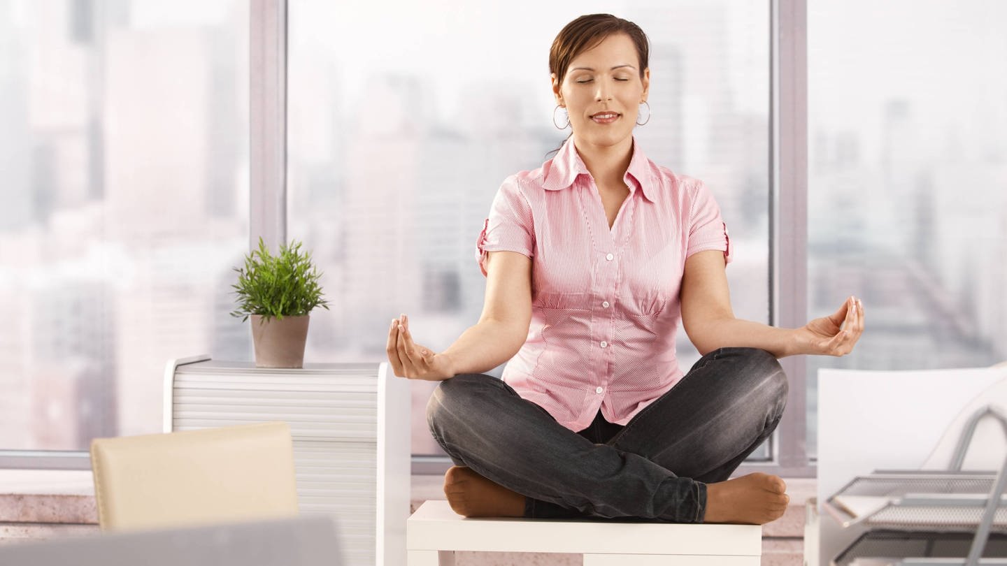 Eine Frau sitzt meditierend auf ihrem Schreibtisch: Stressbewältigung im Job ist wichtig für die eigene Gesundheit (Foto: IMAGO, IMAGO / Shotshop)