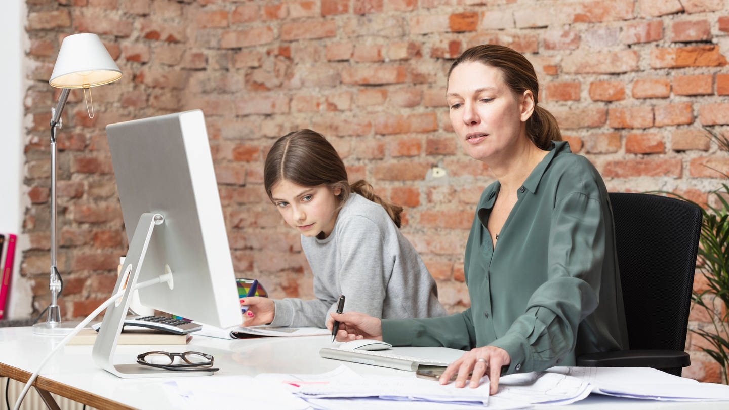 Mutter arbeitet im Homeoffice am Laptop, Tochter macht daneben Hausaufgaben: Eltern leisten 