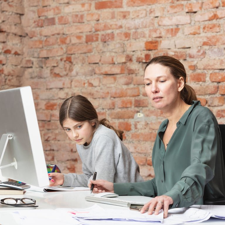 Mutter arbeitet im Homeoffice am Laptop, Tochter macht daneben Hausaufgaben: Eltern leisten "nebenbei" noch die Betreuung der Kinder