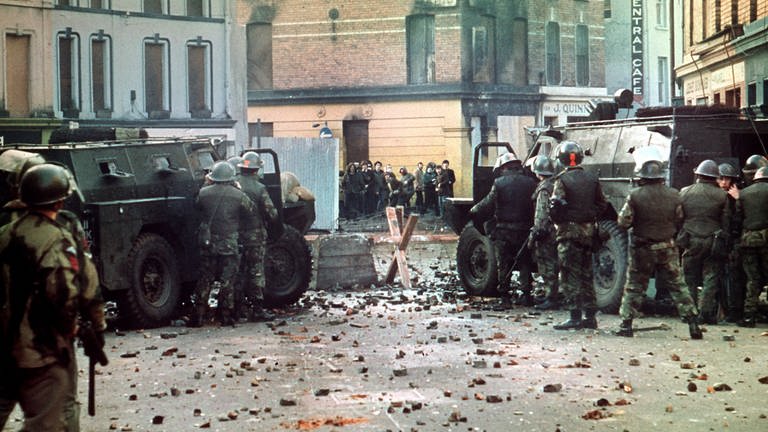 Britische Soldaten mit Panzerwagen stehen auf einer mit Steinen übersäten Straße hinter einer Straßensperre, in einiger Entfernung eine Gruppe von Demonstranten. Am 30. Januar 1972 wurden 13 katholische Demonstranten während einer friedlichen, jedoch verbotenen Kundgebung in der nordirischen Stadt Derry von britischen Fallschirmjägern erschossen. (Foto: dpa Bildfunk, picture alliance/dpa | UPI)