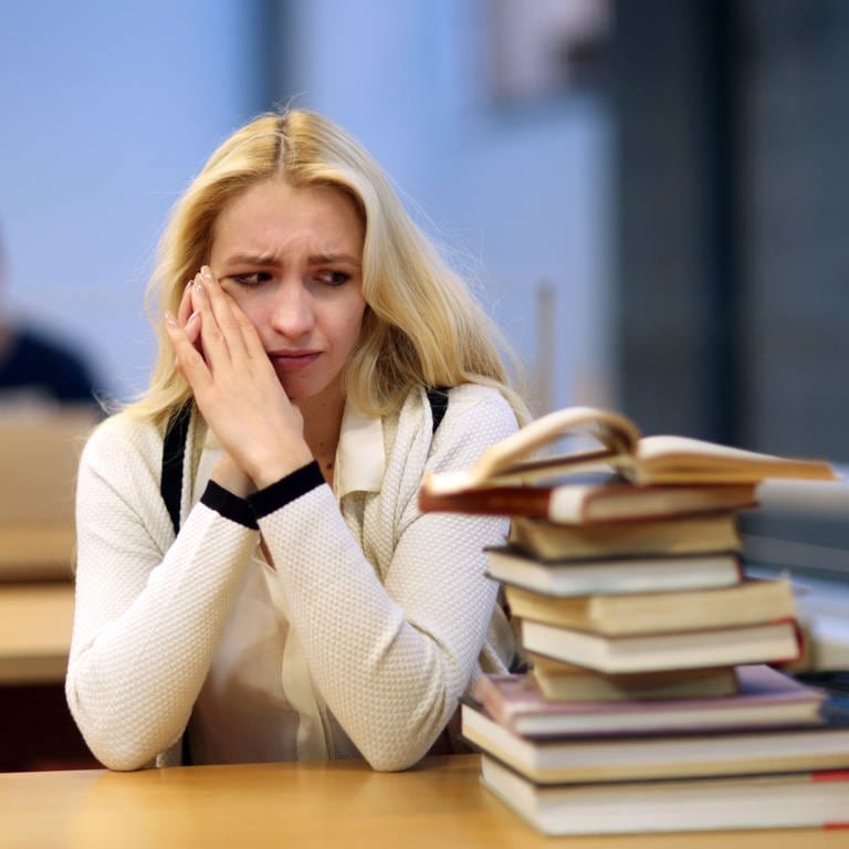 Eine junge Frau sitzt in einer Bibliothek und starrt auf einen Bücherstapel: Vor allem wer selbstständig am Computer arbeitet oder studiert, ist Prokrastination bzw. Aufschieberitis konfrontiert