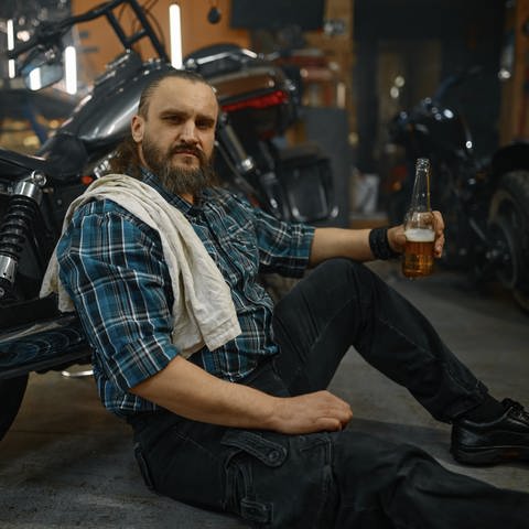 Ein Biker sitzt auf dem Boden und hält eine geöffnete Flasche Bier in der Hand (Foto: IMAGO, IMAGO / Panthermedia)