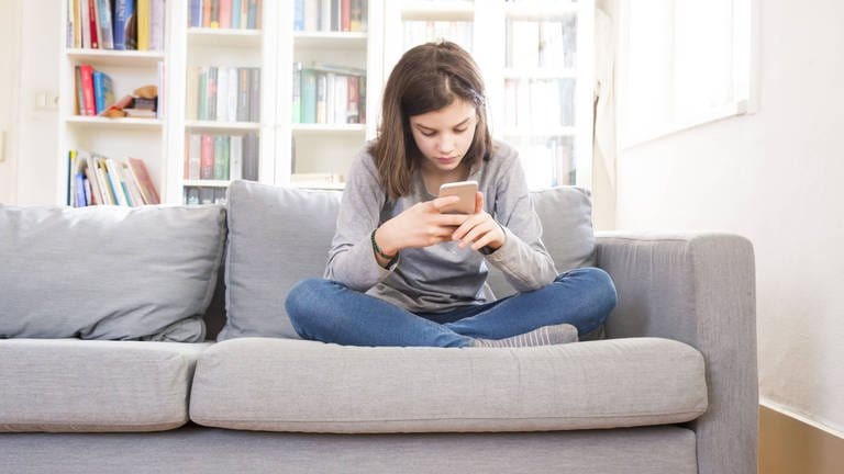 Mädchen sitzt mit Smartphone auf der Couch  (Foto: IMAGO, imago/Westend61)