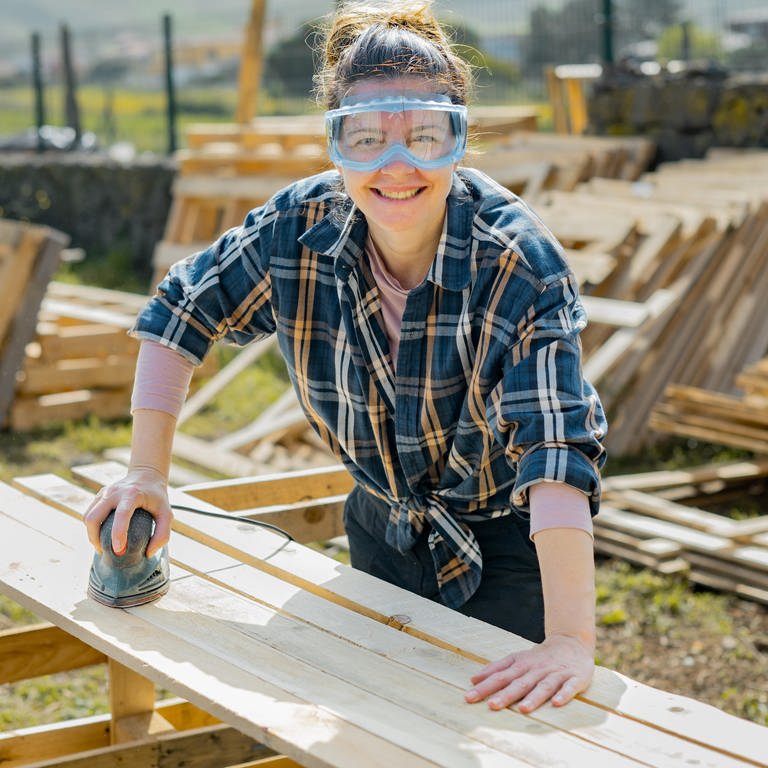 Frau mit Schutzbrille schleift Bretter auf einer Baustelle: Ob auf dem Dachstuhl, der Malerleiter oder am Sicherungskasten – Handwerkerinnen sind immer noch die große Ausnahme. 