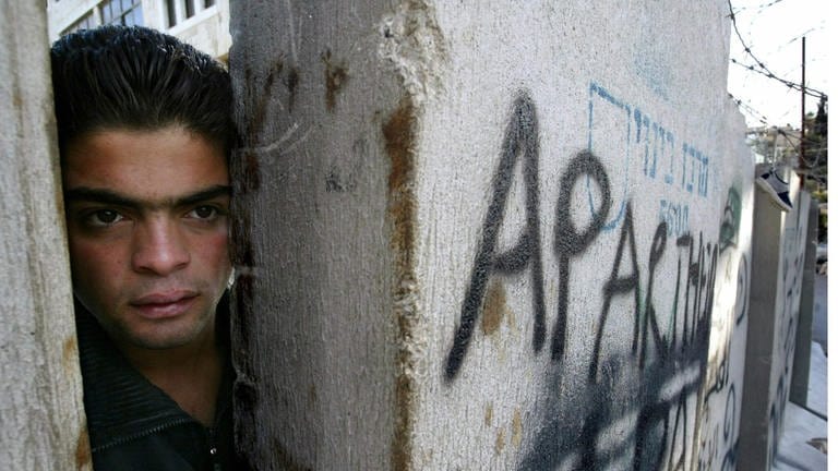 Ein Palästinenser, der auf der Westjordanlandseite des „Trennzauns“ in Abu Dis steht, schaut im November 2003 durch eine Lücke, um zu sehen, ob israelische Polizisten in der Nähe auf der Ost-Jerusalem-Seite der Mauer sind. Auf der Mauer steht "Apartheid".