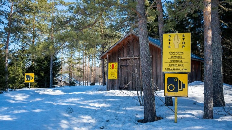 Holzhütte in einem verschneiten Kiefernwald, darin einige gelbe Warnschilder: Grenze zu Russland im finnischen Vaalimaa