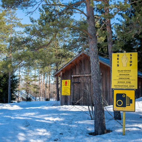 Holzhütte in einem verschneiten Kiefernwald, darin einige gelbe Warnschilder: Grenze zu Russland im finnischen Vaalimaa