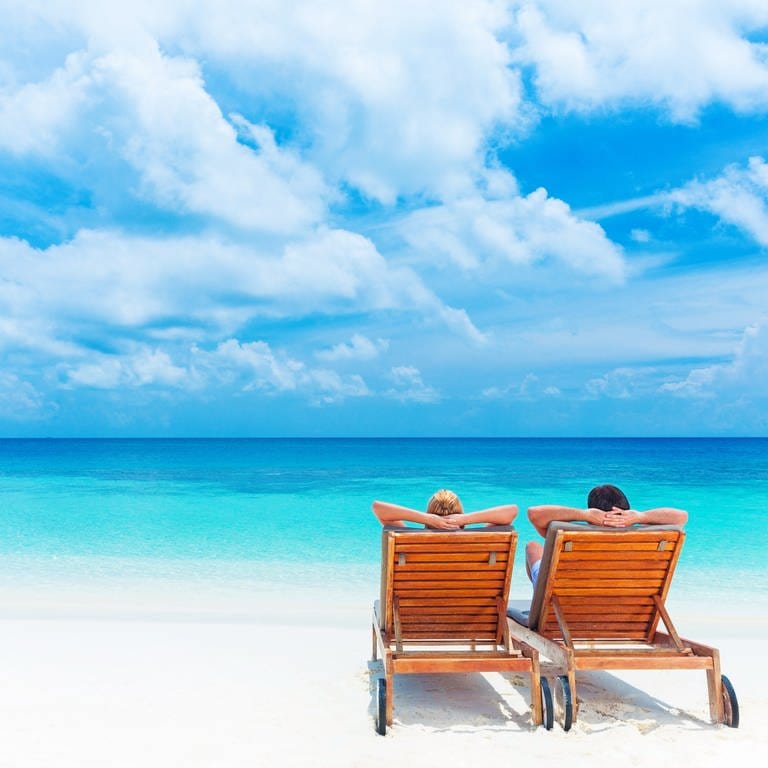 Ein Paar liegt am Strand auf Holzliegen und schaut aufs blaue Meer: Palmen, Sand und blaues Meer: Unser Traum vom Strandparadies ist gerade mal 150 Jahre alt. Der Aufenthalt am Meeresrand galt erst als gefährlich, dann als heilsam. (Foto: Colourbox)