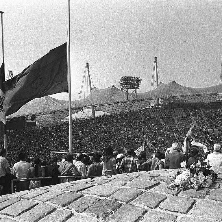 Blumen liegen am 6. September 1972 während des Gedenkgottesdienstes für die elf ermordeten israelischen Teammitglieder am Fuße der olympischen Fackel im Münchner Olympiastadion, eine Fahne weht auf Halbmast. Das Stadion mit 80.000 Sitzplätzen war während der Zeremonie bis auf den letzten Platz gefüllt. (Foto: picture-alliance / Reportdienste, picture alliance / AP | AP)