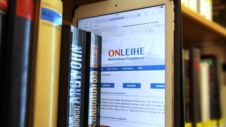 Ein iPad mit der Internetseite der Onleihe Mecklenburg-Vorpommern steckt zwischen den Büchern im Regal einer Bibliothek: Autoren und Verlage sehen ihre Existenz durch die sogenannte Onleihe gefährdet, Bibliotheken beklagen, dass ihnen Bestseller oft erst Monate nach Erscheinen verkauft werden. 