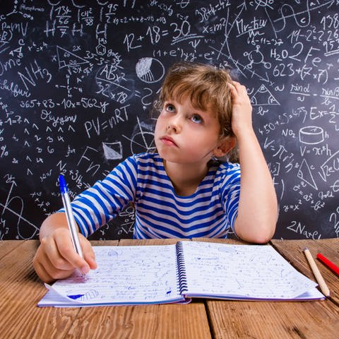 Junge sitzt vor Schultafel mit vielen Matheaufgaben und denkt nach: Schlecht in Mathe – oft liegt das an schlechtem Unterricht, nicht an mangelndem Talent. Gute Mathe-Didaktik setzt auf Verstehen statt auswendig lernen und begeistert für das Fach.