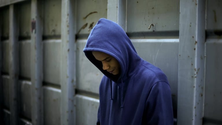 Junge mit blauem Kapuzenshirt lehnt an einem Gitter und schaut nach unten: Etwa 40.000 Minderjährige und junge Menschen in Deutschland haben kein festes Zuhause. Was treibt die Jungendlichen auf die Straße? Wann hilft ihnen der Staat, wo sind Lücken? Und was wünschen sich junge Obdachlose?