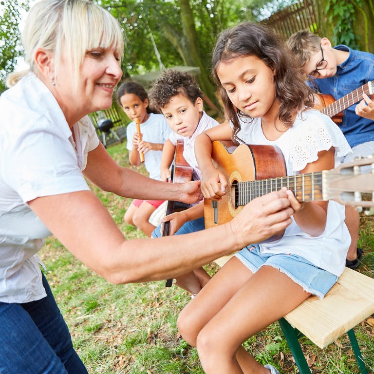 Kinder lernen Gitarre spielen, eine Lehrerin hilft: Durch Musik lernen Kinder und Jugendliche, einander zuzuhören, erleben Gemeinschaft und verstehen die Kultur eines Landes. Dennoch fällt Musikunterricht regelmäßig aus oder wird fachfremd unterrichtet. 