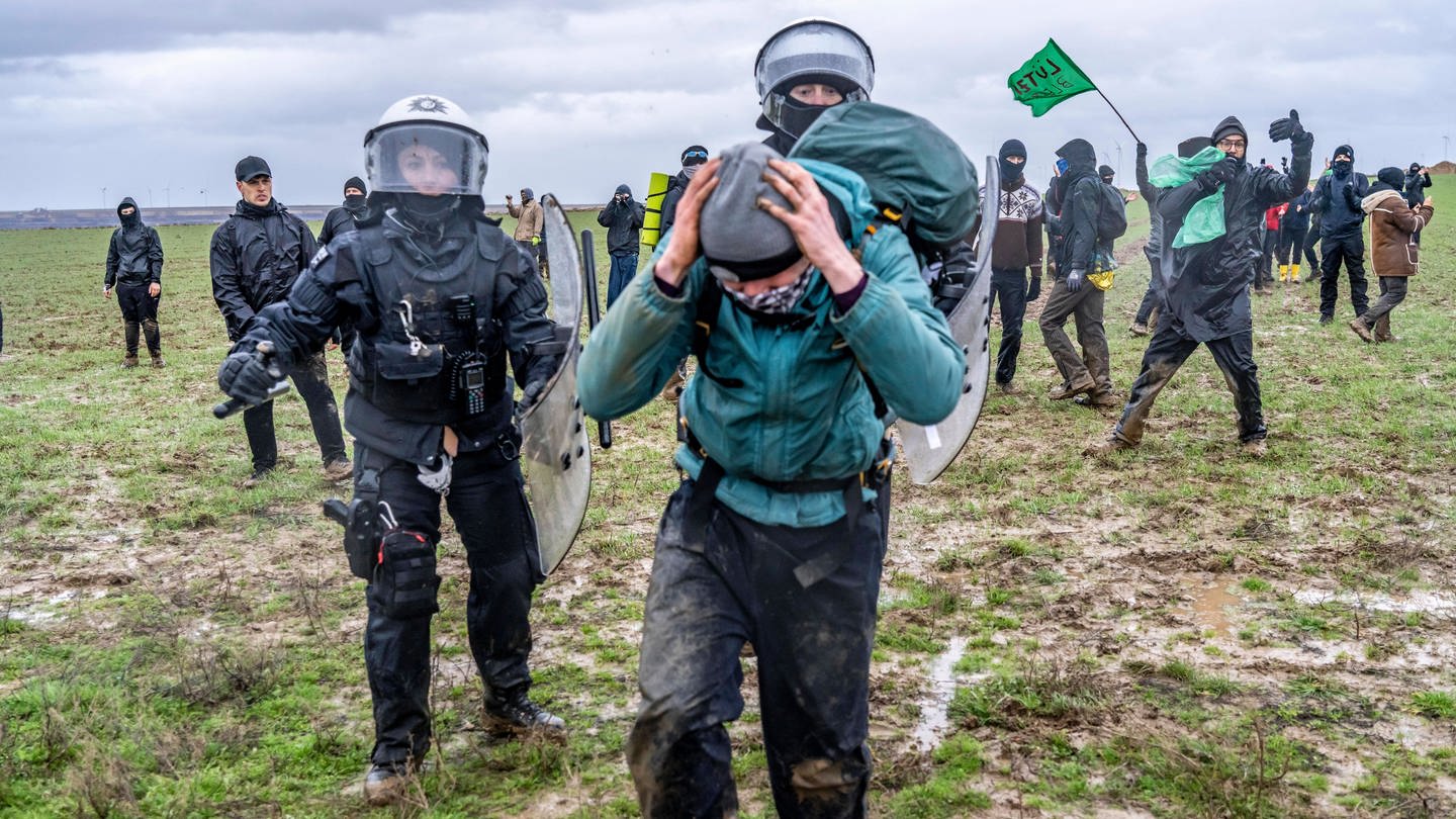 Zwei Polizisten in Schutzkleidung verfolgen einen Demonstranten auf einem Feld bei Lützerath (Foto: IMAGO, Jochen Tack)