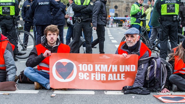 Klima-Aktivisten der "Letzten Generation" haben sich am Karlsplatz in München festgeklebt. Auf einem Transparent steht "100 kmh und 9 € für alle" (Foto: picture-alliance / Reportdienste, picture alliance / ZUMAPRESS.com | Sachelle Babbar)