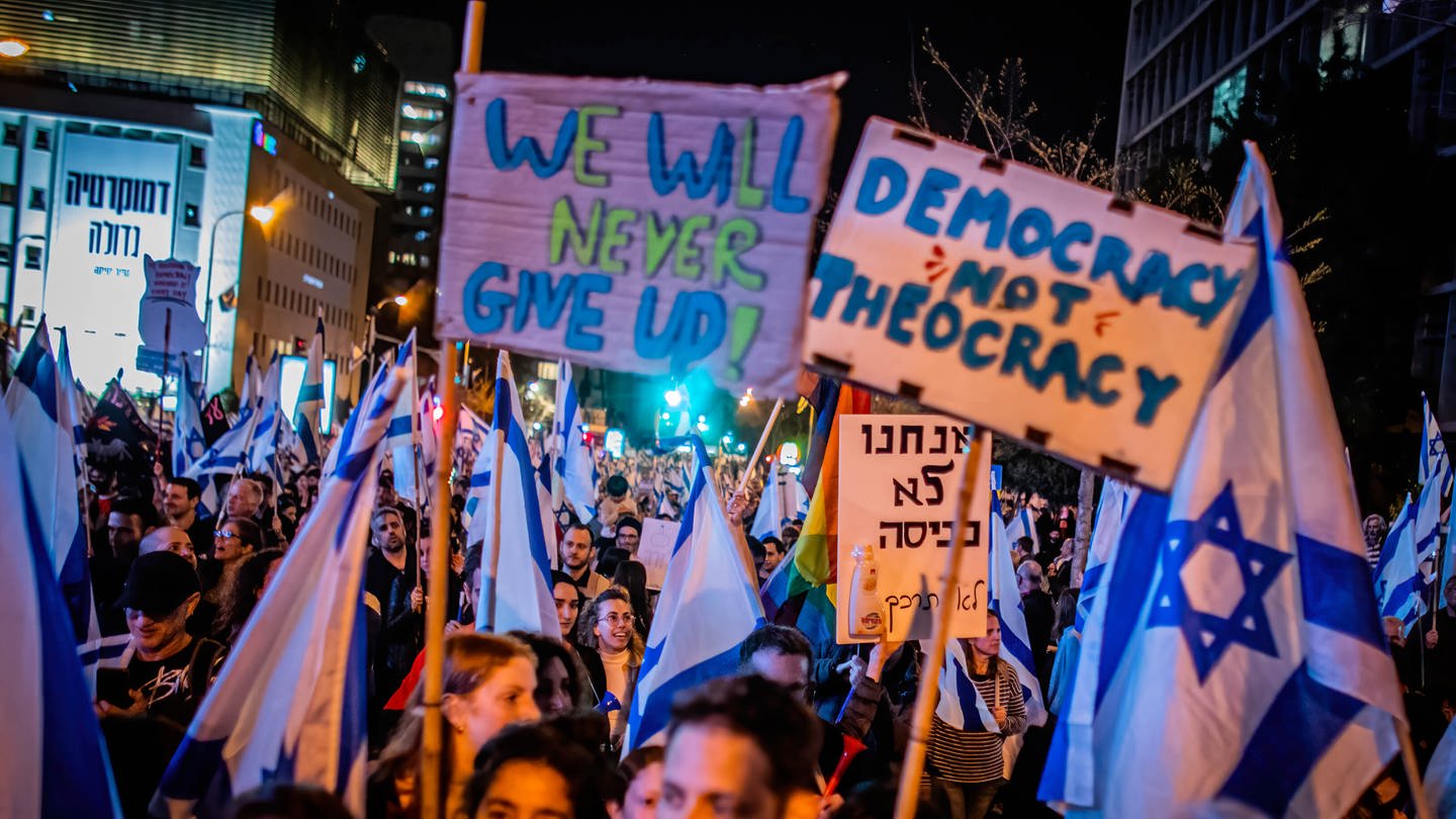 Demonstranten halten am 25. März 2023 in Tel Aviv / Israel Fahnen und Plakate in die Höhe. Auf einem steht 