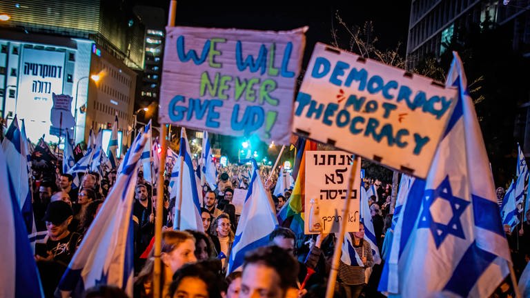 Demonstranten halten am 25. März 2023 in Tel Aviv  Israel Fahnen und Plakate in die Höhe. Auf einem steht "Democracy not Theocracy". Der Protest richtet sich gegen Pläne der Regierung, das Justizsystem umzustrukturieren. 