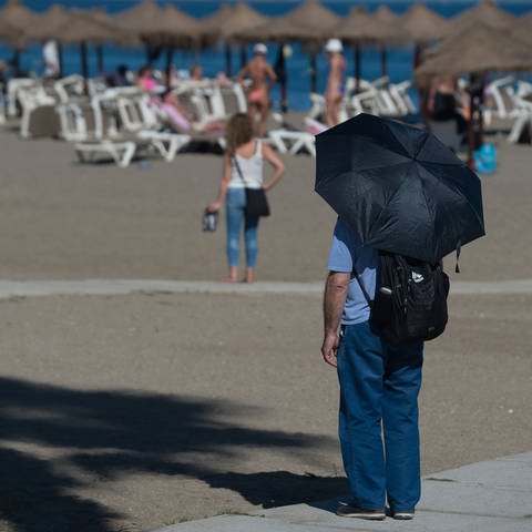 Ein Mann steht mit einem Schirm am Strand, um sich vor der Sonne zu schützen (Foto: IMAGO, IMAGO / ZUMA Wire)