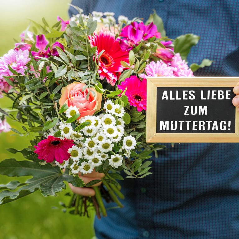 Ein Mann hält einen Blumenstrauß und eine Tafel in der Hand mit Aufschrift: Alles Liebe zum Muttertag. (Foto: picture-alliance / Reportdienste, picture alliance / CHROMORANGE | Michael Bihlmayer)