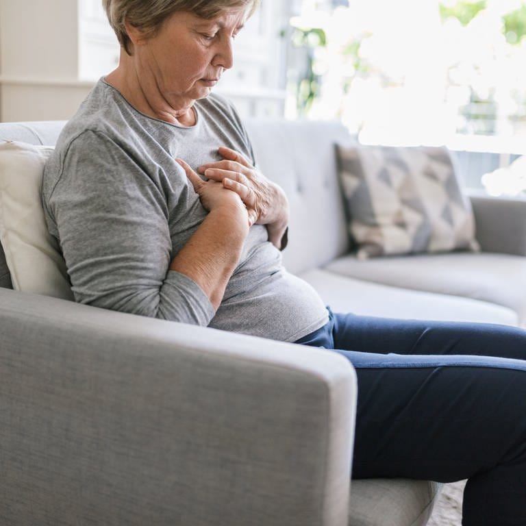 Eine Frau sitzt auf einem grauen Sofa und greift sich an die Brust: Der weibliche Herzinfarkt wird oft später erkannt und später behandelt. 