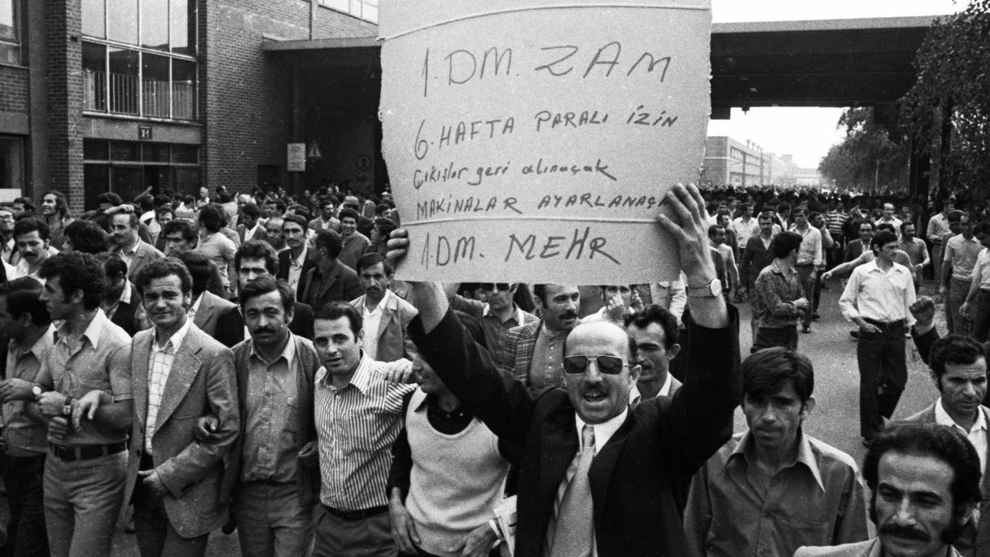 Der Streik bei den Ford-Werken, hier am 27. bis 29. 8. 1973 in Köln, war ein Spontanstreik, also ein wilder Streik. Die Serie von Streiks wurde u. von türkischen Migranten organisiert. Ursache war u. a. die starke Inflation. Auf dem Schild in türkischer Sprache, das einer der Männer in die Höhe hält, steht in der letzten Zeile auf Deutsch 