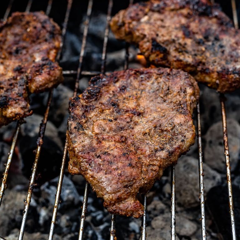 Auf dem Grill liegen drei Fleischstücke. (Foto: IMAGO, IMAGO / Wirestock)