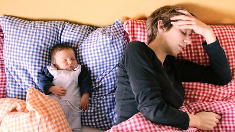 Mutter und Neugeborenes liegen im Bett, die Mutter liegt weg gedreht vom Baby und hält sich ihre Hand an die Stirn.