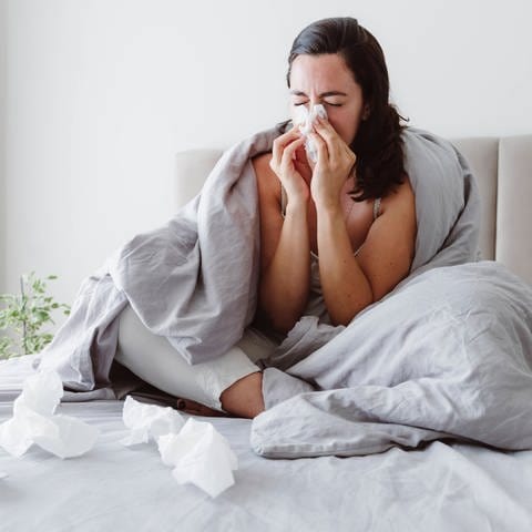 Eine Frau sitzt krank auf dem Bett und putzt sich die Nase.