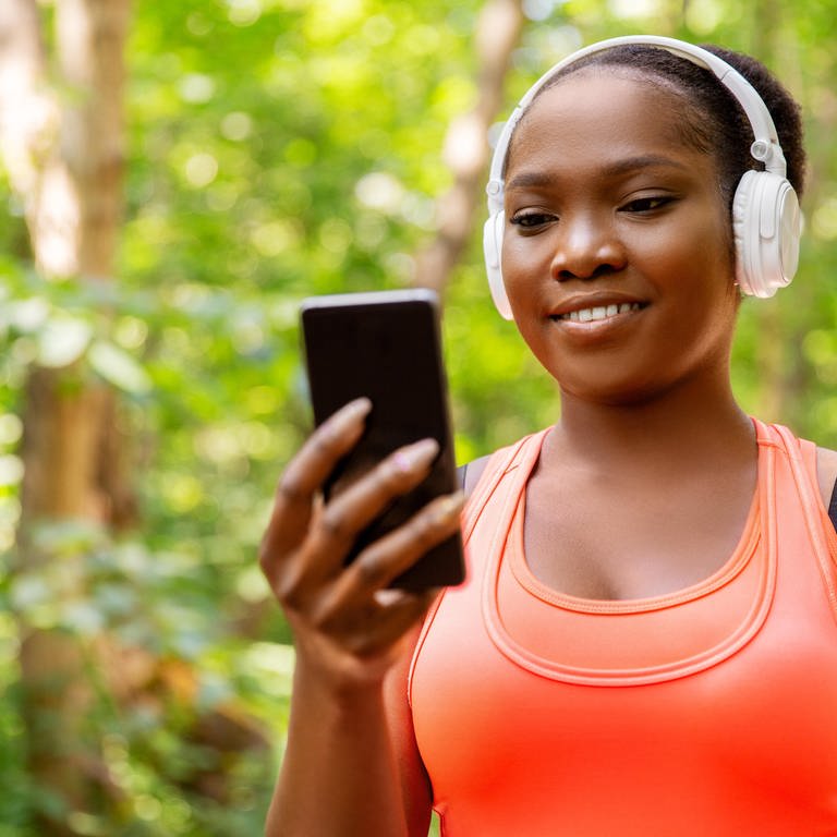 Junge Frau mit Kopfhörer und Smartphone geht durch den Wald: Das Radio der Zukunft soll die Höererinnen und Hörer unterwegs mit relevanten Informationen versorgen und mittel KI mit ihnen in Dialog treten können.