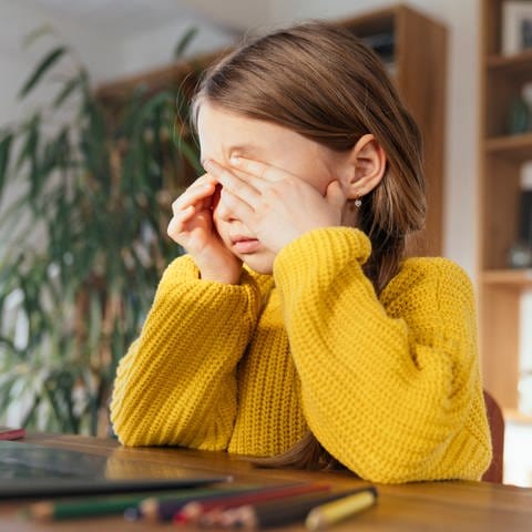 Ein Mädchen reibt sich die Augen, während es zu Hause vor dem Laptop sitzt.